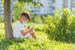 un niño en un médico máscara se sienta en el césped y mira en el teléfono dibujos animados en el verano a puesta de sol. niño con un móvil teléfono en su manos. prevención en contra coronavirus covid-19 durante un pandemia foto