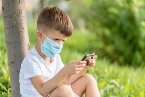 un niño en un médico máscara se sienta en el césped y mira en el teléfono dibujos animados en el verano a puesta de sol. niño con un móvil teléfono en su manos. prevención en contra coronavirus covid-19 durante un pandemia foto