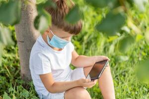 un niño en un médico máscara se sienta en el césped y mira en el teléfono dibujos animados en el verano a puesta de sol. niño con un móvil teléfono en su manos. prevención en contra coronavirus covid-19 durante un pandemia foto