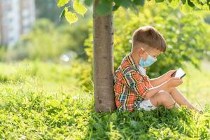 un niño en un médico máscara se sienta en el césped y mira en el teléfono dibujos animados en el verano a puesta de sol. niño con un móvil teléfono en su manos. prevención en contra coronavirus covid-19 durante un pandemia foto