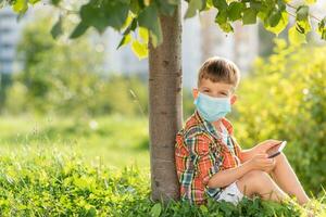 un niño en un médico máscara se sienta en el césped y mira en el teléfono dibujos animados en el verano a puesta de sol. niño con un móvil teléfono en su manos. prevención en contra coronavirus covid-19 durante un pandemia foto