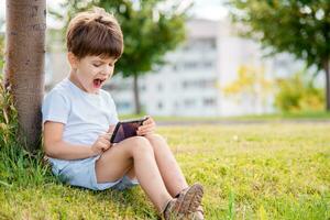 Cheerful child sitting on the grass looks cartoons in the phone in the summer at sunset. Cute boy having fun in nature photo