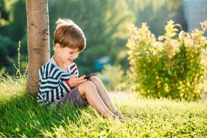 Cheerful boy sitting on the grass looks cartoons in the phone in the summer at sunset. Cute baby having fun in nature photo