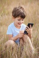 A beautiful child sitting on the grass speaks by phone in the summer at sunset. Boy communicates on mobile photo