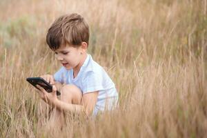 un hermosa niño sentado en el césped habla por teléfono en el verano a puesta de sol. chico comunica en móvil foto