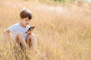 alegre niño sentado en el césped mira dibujos animados en el teléfono en el verano a puesta de sol. linda chico teniendo divertido en naturaleza foto