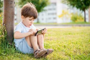 alegre niño sentado en el césped mira dibujos animados en el teléfono en el verano a puesta de sol. linda chico teniendo divertido en naturaleza foto