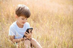 alegre niño sentado en el césped mira dibujos animados en el teléfono en el verano a puesta de sol. linda chico teniendo divertido en naturaleza foto