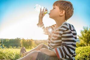 linda chico sentado en el césped bebidas agua desde un botella en el verano a puesta de sol. niño apaga sed en un caliente día foto