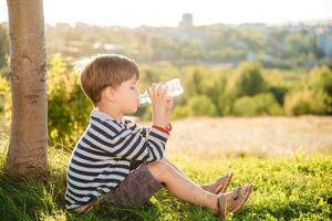 Cute boy sitting on the grass drinks water from a bottle in the summer at sunset. Child quenches thirst on a hot day photo