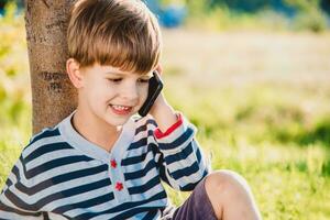 un hermosa niño sentado en el césped habla por teléfono en el verano a puesta de sol. chico comunica en móvil foto