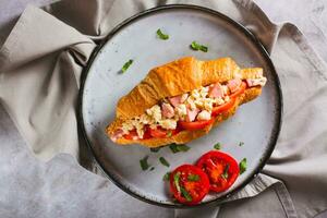 Close up of croissant sandwich with scrambled eggs, sausages, tomatoes and herbs on a plate top view photo