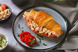 Delicious croissant sandwich with scrambled eggs, sausages and tomatoes on a plate photo