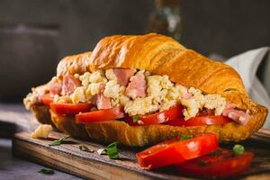 Close up of croissant sandwich with scrambled eggs, sausages and tomatoes on a board photo