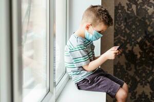 A cute child in a medical mask sits at home in quarantine by the window with a phone in his hands and looks into it. Prevention of coronavirus and Covid - 19 photo