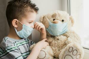 a small child in a mask sits at home in quarantine and looks out the window in a place with a teddy bear. Prevention of Coronavirus and Covid - 19. Concept photo