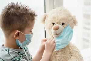 A small child helps a teddy bear put on a medical mask at home in quarantine. Prevention of coronavirus and Covid - 19. Concept photo