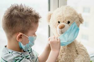 A small child helps a teddy bear put on a medical mask at home in quarantine. Prevention of coronavirus and Covid - 19. Concept photo