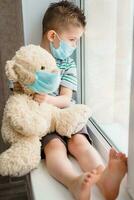 a small child in a mask sits at home in quarantine and looks out the window in a place with a teddy bear. Prevention of Coronavirus and Covid - 19. Concept photo