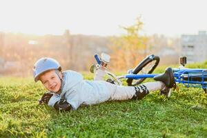 un pequeño niño en un casco y proteccion cayó desde un bicicleta sobre el césped y estaba no lesionado foto