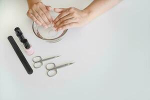 Hands of woman are in bath with warm water with useful substances. Nail file, scissors and bottles of varnish are next on table, copy space. Concept of nail and cuticle and hand skin care photo