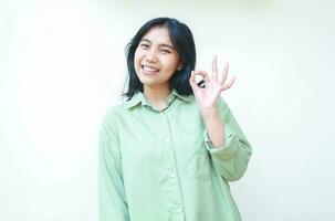 pleased satisfied asian woman with dark hair showing ok sign excelent symbols wearing green over size shirt smiling excited at camera standing over isolated white background photo