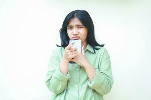 portrait of unhappy sad asian woman looking at camera holding smartphone on chin wearing green clothes showing worried face expression standing isolated white background photo