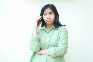 thoughtful asian woman wearing over sized green clothes showing sad face expression while talking on smartphone with folding arms isolated on white background photo