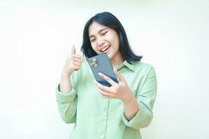 playful confident asian woman with dark hair wearing green over size shirt showing thumbs up looking at smartphone with joy recommend good product standing isolated white background photo