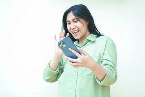 confident ecstatic success asian woman dark haired raising palms say hi gesture to smartphone hold on hands wearing green over size shirt standing isolated white background, welcoming expression photo