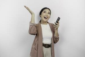 Excited Asian woman employee wearing cardigan, holding her phone, and pointing at the copy space above her, isolated by white background photo