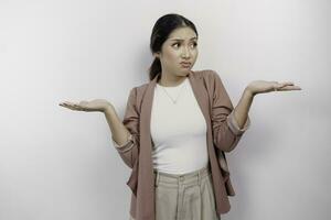 A thoughtful Asian woman employee wearing a cardigan is pointing to copy space beside her, looks so confused between choices, isolated by a white background photo