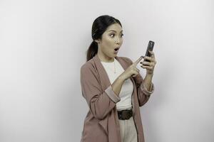 conmocionado joven asiático mujer empleado vistiendo cárdigan es participación su teléfono, aislado por blanco antecedentes foto