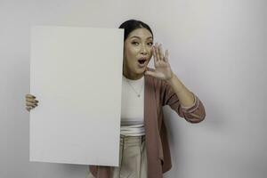 Young beautiful Asian woman employee wearing cardigan is shouting and screaming loud with a hand on her mouth while holding empty blank board, isolated by white background. photo