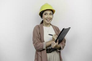 A thoughtful young Asian woman employee wearing safety helmet while holding a clipboard and looking aside to copy space, isolated by white background. photo