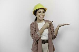 Excited Asian woman employee wearing safety helmet pointing at the copy space beside her, isolated by white background photo