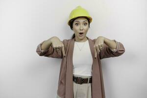 Shocked Asian woman employee wearing safety helmet pointing at the copy space below her, isolated by a white background photo