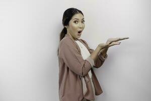 Shocked Asian woman employee wearing a cardigan pointing at the copy space beside her, isolated by a white background photo