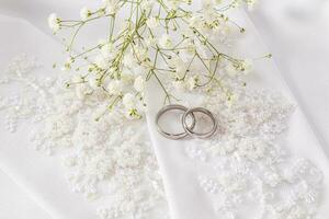 Two white gold wedding rings lie on the bride's chic wedding gloves. White satin background. Wedding accessories concept. photo