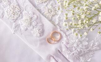 Two gold wedding rings lie on the bride's chic beaded gloves embroidered with beads and sequins. Top view. Satin white background. photo