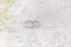 Two white gold wedding rings lie on the bride's beaded and pearled gloves. white satin background. with a branch of gypsophila. Wedding concept. photo