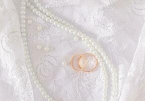 Two gold wedding rings lie on a luxurious vintage part of the bride's wedding dress with a string of pearl beads. Wedding concept. Top view photo