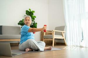 Asian senior woman lifting dumbbell for exercise and workout at home. Active mature woman doing stretching exercise in living room. Exercise Active and healthy for older, elder, and senior concept. photo