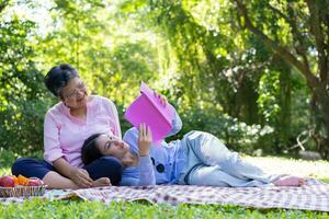 Asian daughter Sleeping on mother lap and reading book on picnic mat in the park. A happy senior woman talks with her daughter. Concept of healthcare and elderly caregiver support service. photo