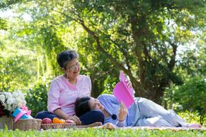 Asian daughter Sleeping on mother lap and reading book on picnic mat in the park. A happy senior woman talks with her daughter. Concept of healthcare and elderly caregiver support service. photo