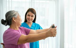 Happy elderly female exercising with rubber expander with Young physiotherapist for recovery arm muscles in a physiotherapy center. Concept of happy retirement with care from a caregiver and health. photo