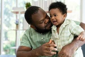 Happy african american man play with little infant toddler child in living room sofa. Parent and little kid relaxing at home. Parents and toddler child girl enjoying spending time at home. Childcare photo