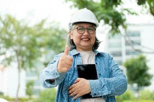 Happy elderly Asian professional engineer wears a helmet and checks the blueprint on a tablet on the construction site after the contractor and architect finish renovation the house and building photo