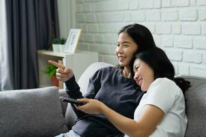 Beautiful smiling LGBTQ Asian lesbian couple holding remote control sitting on sofa at home. photo