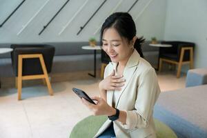hermosa joven asiático mujer usos teléfono inteligente en café comercio, recibe bueno Noticias correo electrónico mensaje en teléfono inteligente foto